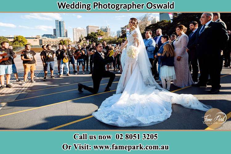 Photo of the Groom on his knee taking the hand of his Bride witnessing by the crowd on the street Oswald NSW 2321